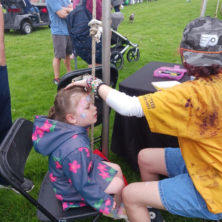 Bex in her element face-painting a young girl in a blue rain-poncho with a pink floral pattern